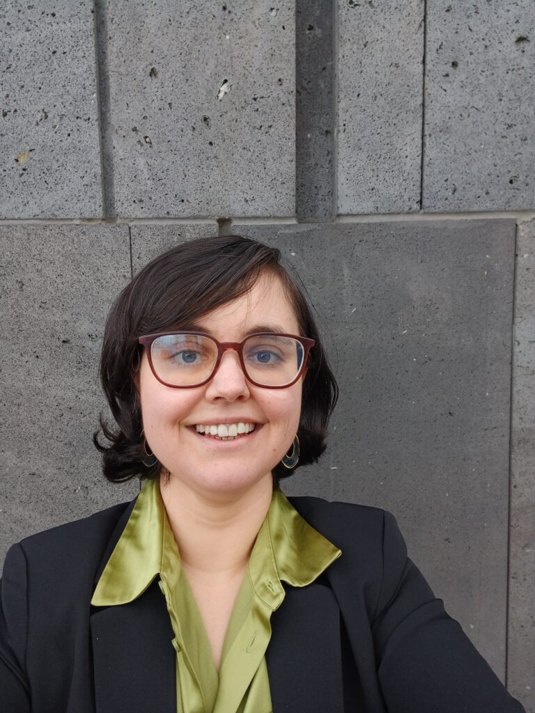 Nahaufnahme einer weiblichen Person mit schwarzen Haaren und Brille, die freundlich in die Kamera lächelt. Sue steht vor einer grauen Betonwand und trägt einen schwarzen Blazer und eine grüne Bluse.