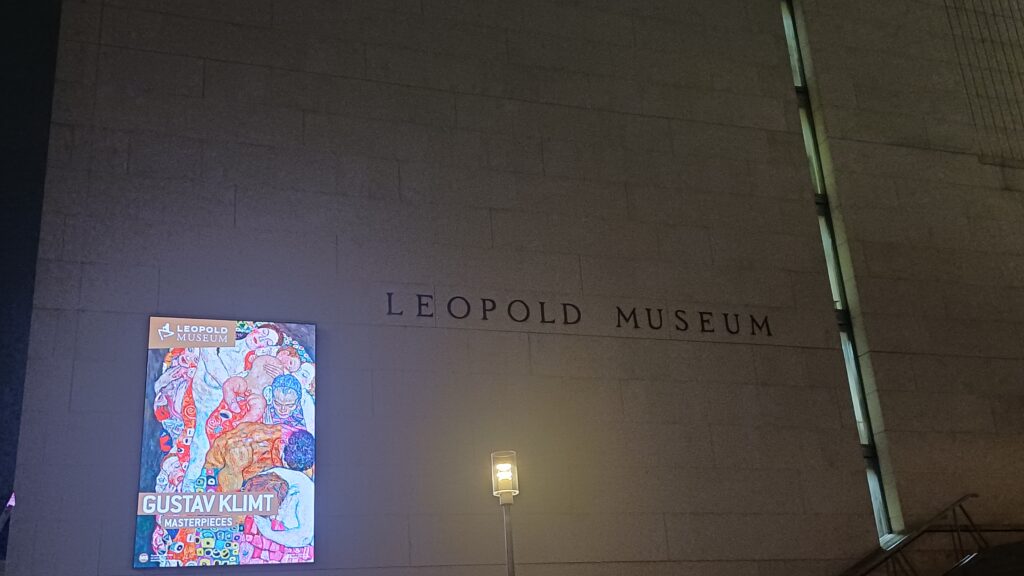 Aufnahme eines Betonbaus mit der Aufschrift Leopold Museum, unter der eine Straßenlaterne brennt. In der linken Bildhälfte ist ein hochkantiger Screen mit einem Bild von Gustav Klimt, das bunte Figuren zeigt, die übereinander liegen.