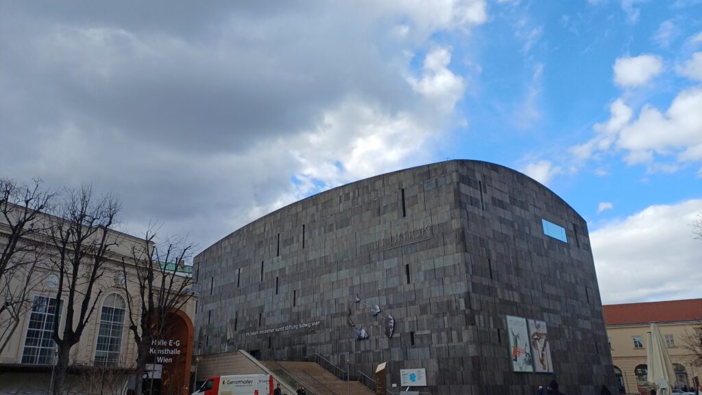 Außenaufnahme eines Flachbaus aus Beton vor blauem Himmel mit Wolken. Neben dem Betongebäude ist ein Stiegenaufgang, daneben Umrisse von Backsteinbauten mit bodentiefen Fenstern. Davor sind Laubbäume ohne Blätter