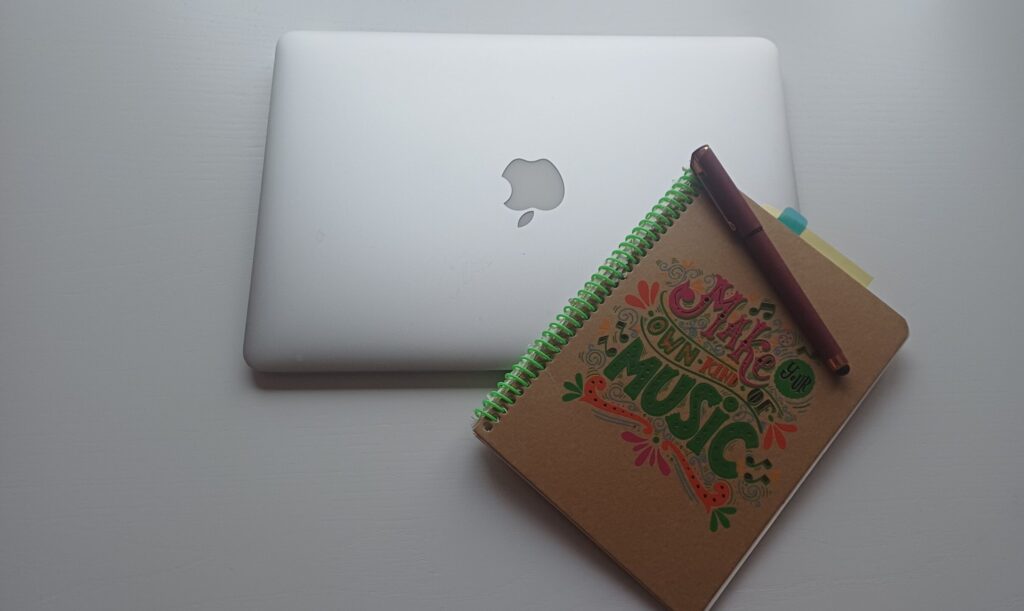 Nahaufnahme eines silbernen, zugeklappten Laptops mit AppleLogo. auf einer weißen Fläche. In der rechten unteren Ecke ist ein Spiral-Notizbuch platziert auf dem die Aufschrift "Make your own kind of music" steht. Darauf liegt ein weinroter Kugelschreiber mit Kappe.