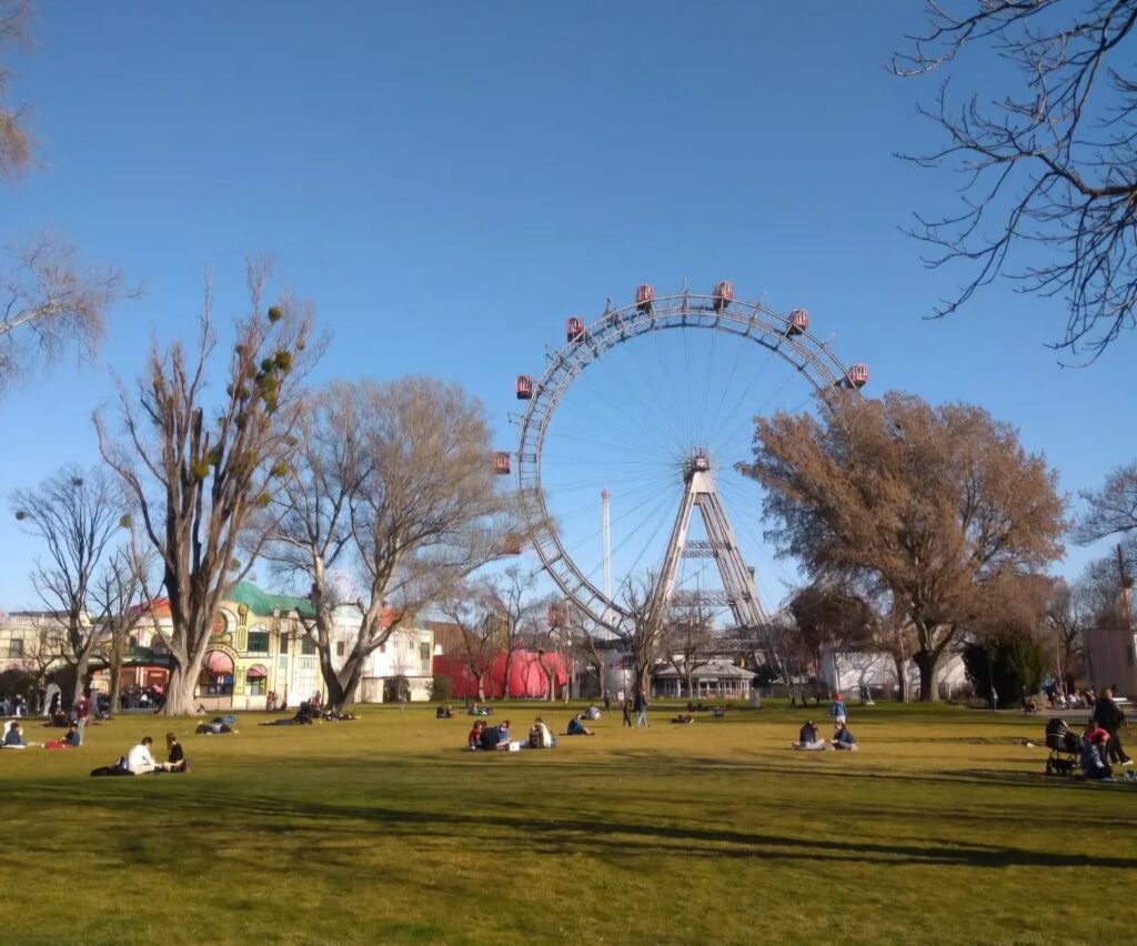 Aufnahme des Wiener Prater bei blauem Himmel. Im Fokus ist das Riesenrad mit roten Gondeln, links davor niedrige Gebäude. Davor ist eine grüne Wiese mit Nadelbäumen und Menschen, die m Gras sitzen.