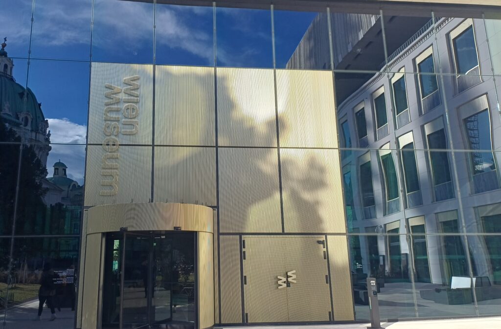 Front aus rechteckigen Spiegeln, in denen die Kuppel einer Kirche, eine Fensterfront und der blaue Himmel mit Wolken. Im Zentrum ist eine beige Drehtür, deren Umrahmung aus beige-bemalten Fenstern besteht und links die Aufschrift Wien Museum enthält.