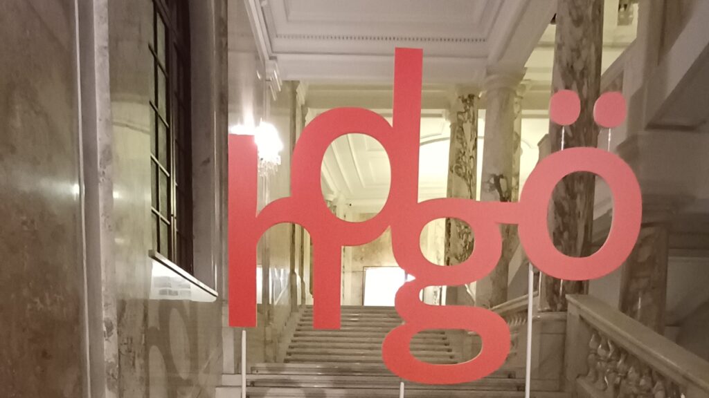 Zu sehen sind rote Aussteller die die Buchtaben hdgö zeigen. Im Hintergrundsind hell beleuchtete Marmortreppen mit einem Geländer und drei marmorierte Säulen