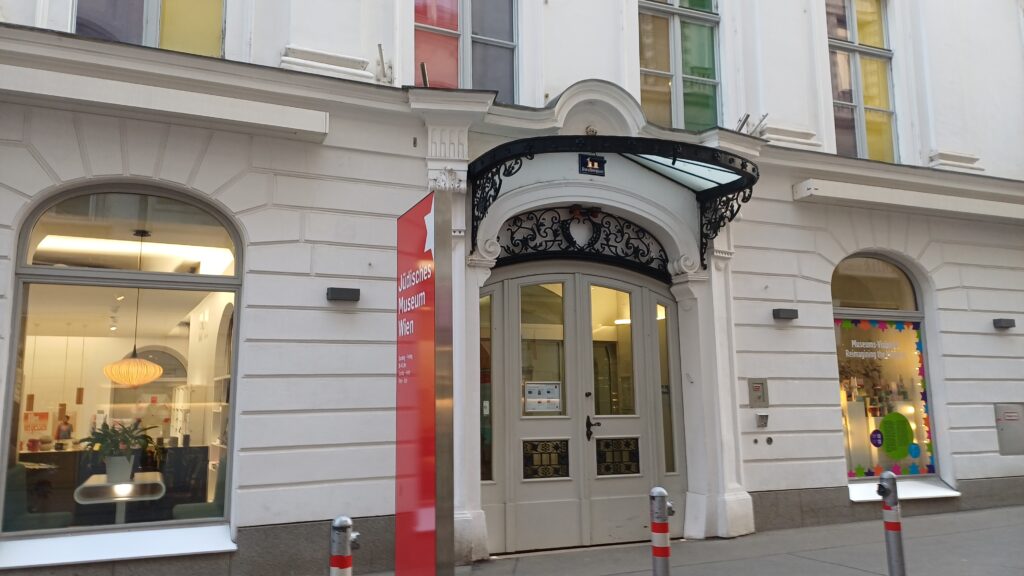 Haus mit weißer Fassade und einem Eingang mit Glastür und einer schwarzen Verzierung und bodentiefen Fenstern. Links vom Eingang ein rotes Roll-up mit weißem Stern und der Aufschrift Jüdisches Museum Wien