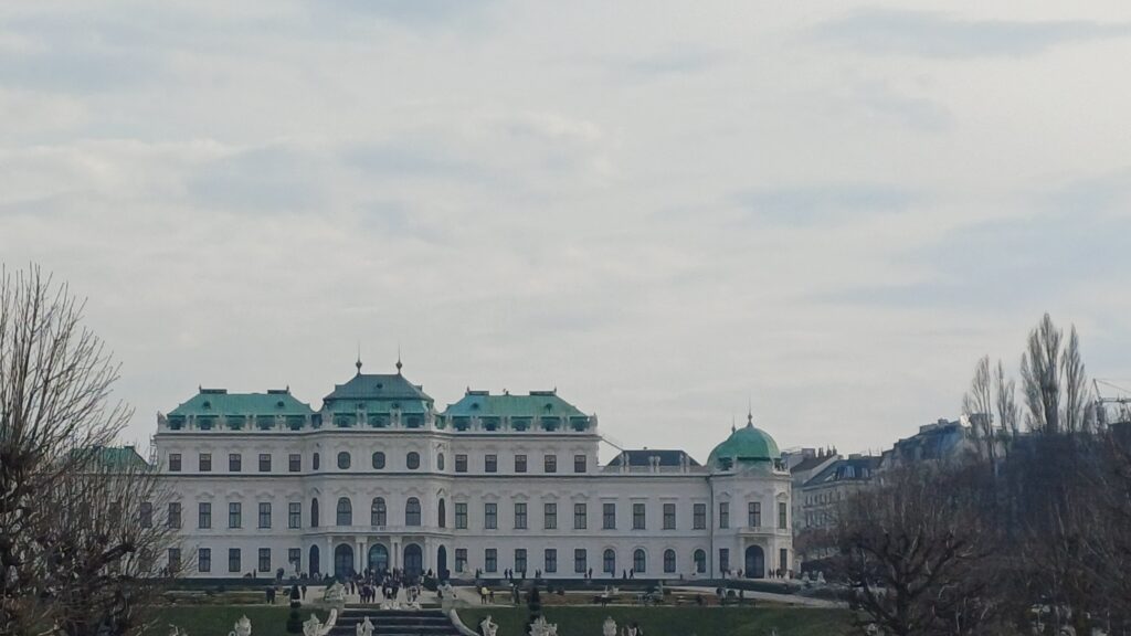Aufnahme des Oberen Belvedere in Wien bei wolkigem Himmel- Im Vordergrund sieht man eine Grünfläche, eine Treppe und Personen, die vor dem Gebäude stehen