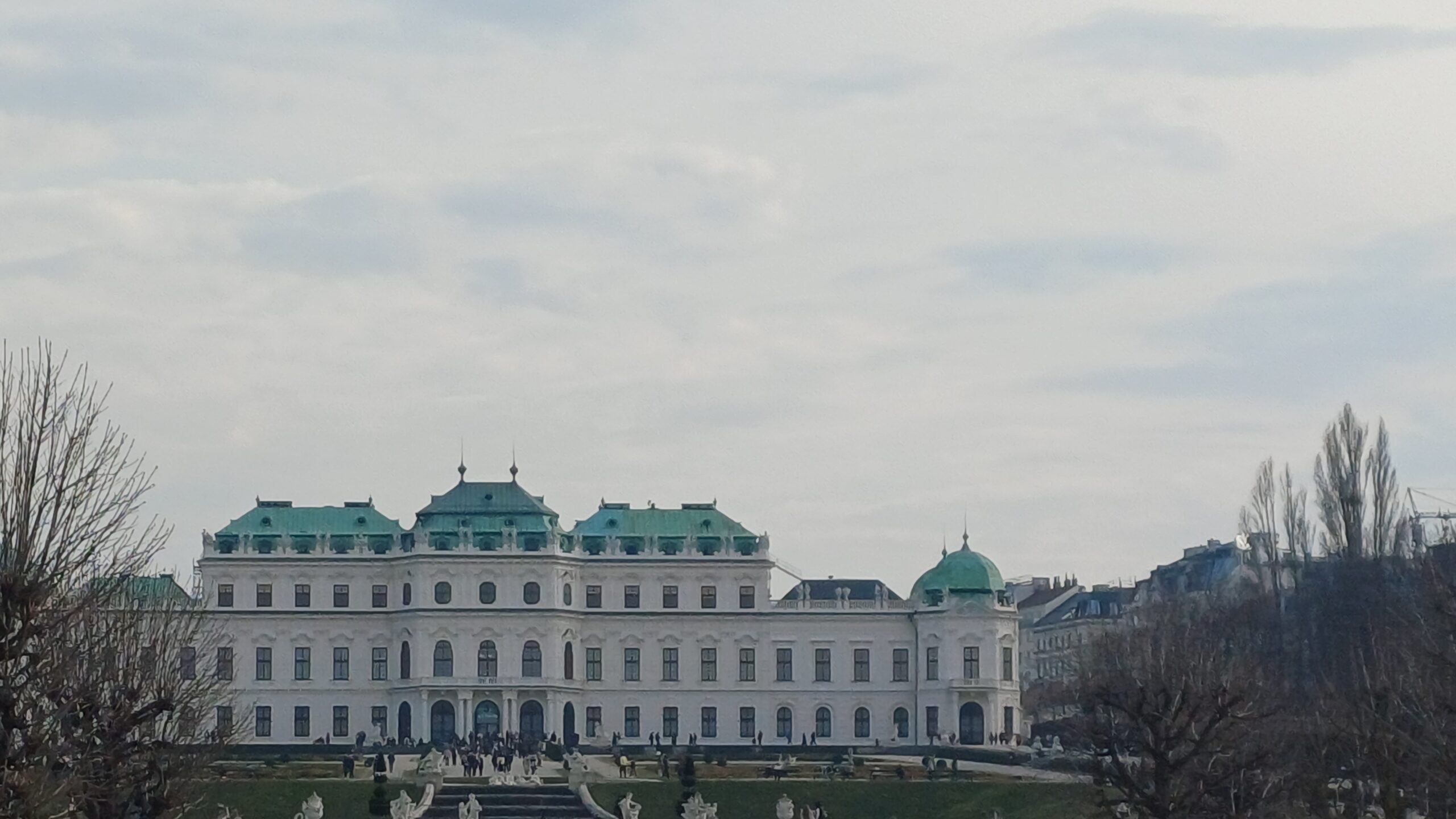 Aufnahme des Oberen Belvedere in Wien bei wolkigem Himmel- Im Vordergrund sieht man eine Grünfläche, eine Treppe und Personen, die vor dem Gebäude stehen