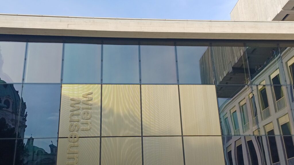 Rückseite eines Gebäudes mit Flachdach, dessen Fassade aus rechteckigen Spiegeln besteht. Sechs rechteckige Elemente mit der vertikalen Aufschrift Wien Museum in beige sind in der Mitte. In den Spiegeln reflektieren Umrisse der Karlskirche