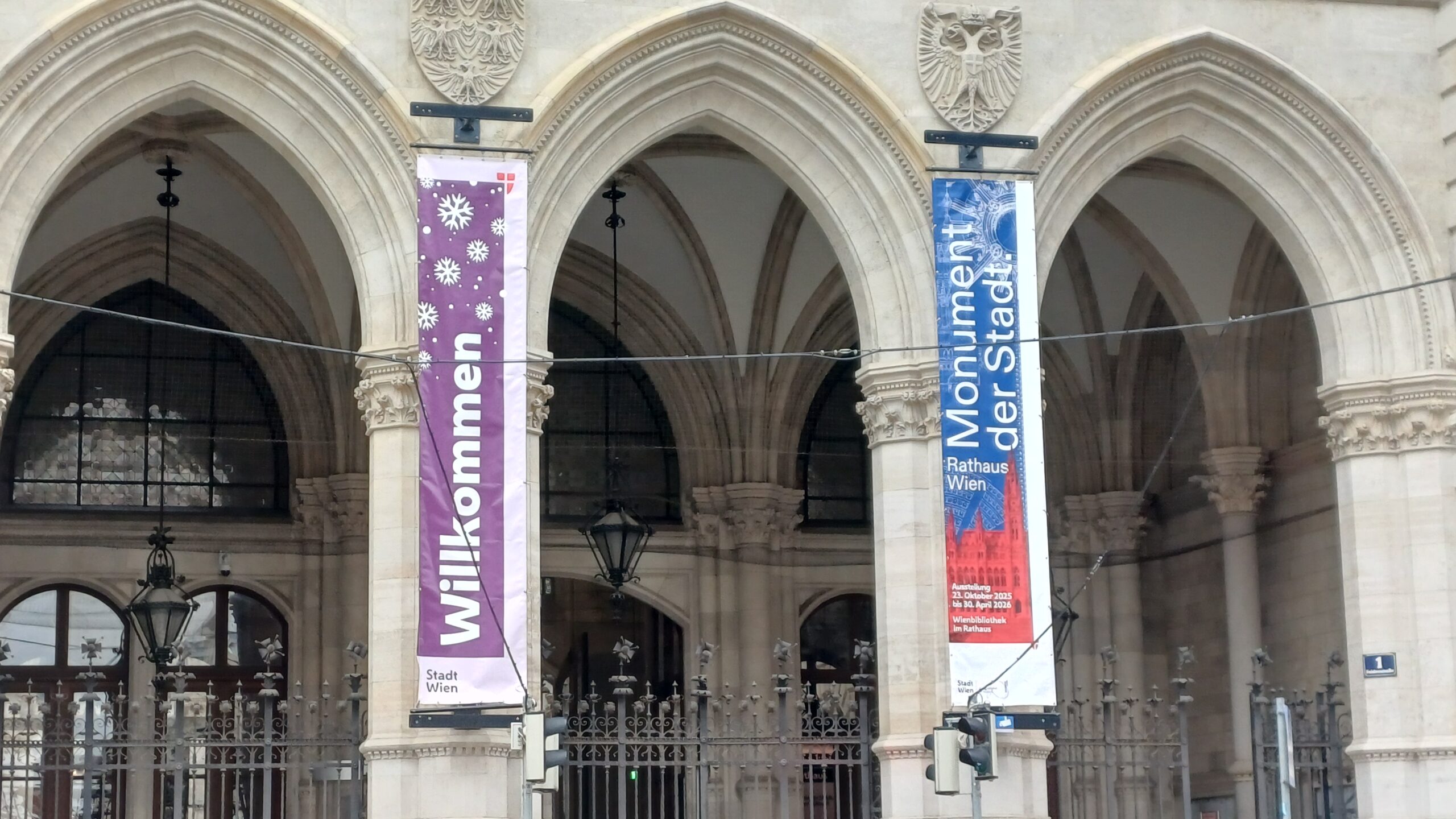 Aufnahme von Arkaden aus hellen Stein, dahinter ein dekorierter Stahlzaun.. Auf den Arkaden sind zwei Stoffbanner vertikal montiert: Auf dem linken steht auf violettem Hintergrund Willkommen in weißer Schrift, auf dem rechten steht Monumente der Stadt in weiß mit blauem Hintergrund. Vor den Arkaden ist ein hängendes Alpensystem montiert.