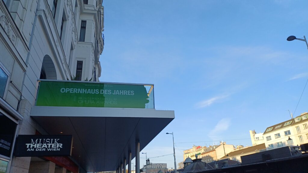 Seitliche Aufnahme des Eingangsbereich eines Backsteinhauses in Form eines Vorbaus auf Säulen eines Backsteinhauses. Auf dem Vorbau ist ein Zaun aus Glasplatten montiert, der auf der sichtbaren Seite die Aufschrift Opernhaus des Jahres in weiß auf grünem Hintergrund enthält Darunter ist ein schwarzes Schild mit der weißen Aufschrift Musik Theater an der Wien montiert.