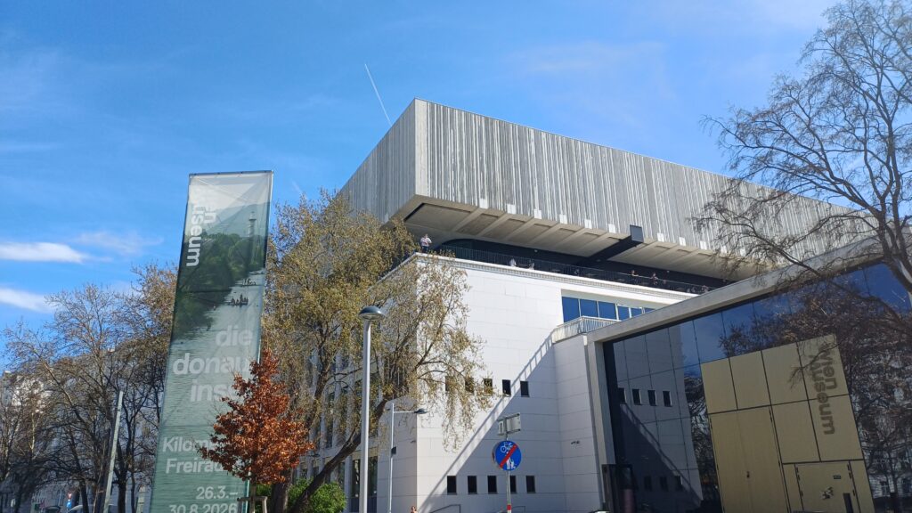 Aufnahme eines Betonbaus mit Flachdach bei blauem Himmel. Der Eingangsbereich des Gebäudes ist verglast und trägt die Aufschrift Wien Museum. Links neben dem Gebäude ist ein Banner mit der Aufschrift Die Donauinsel. ringsherum sind Laubbäume