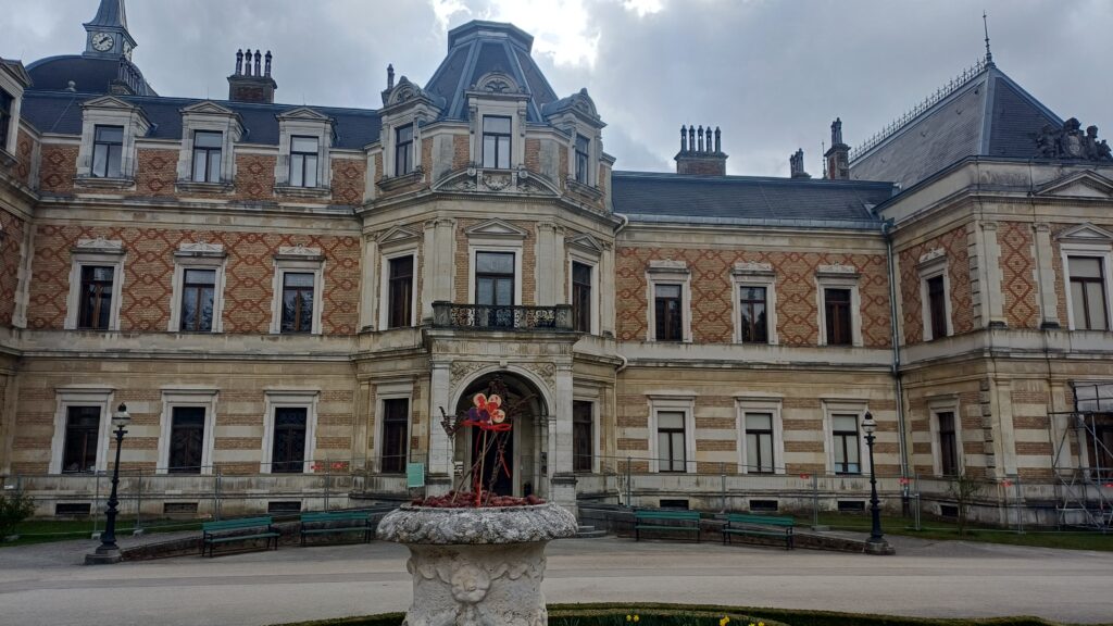 Aufnahme eines zweistöckigen Baus mit einer gelb-orangen Fassade und einem grauen Dach. Der Eingangsbereich und die Seitenschiffe sind vorgelagert. Vor dem Bau ist ein Marmorbrunnen