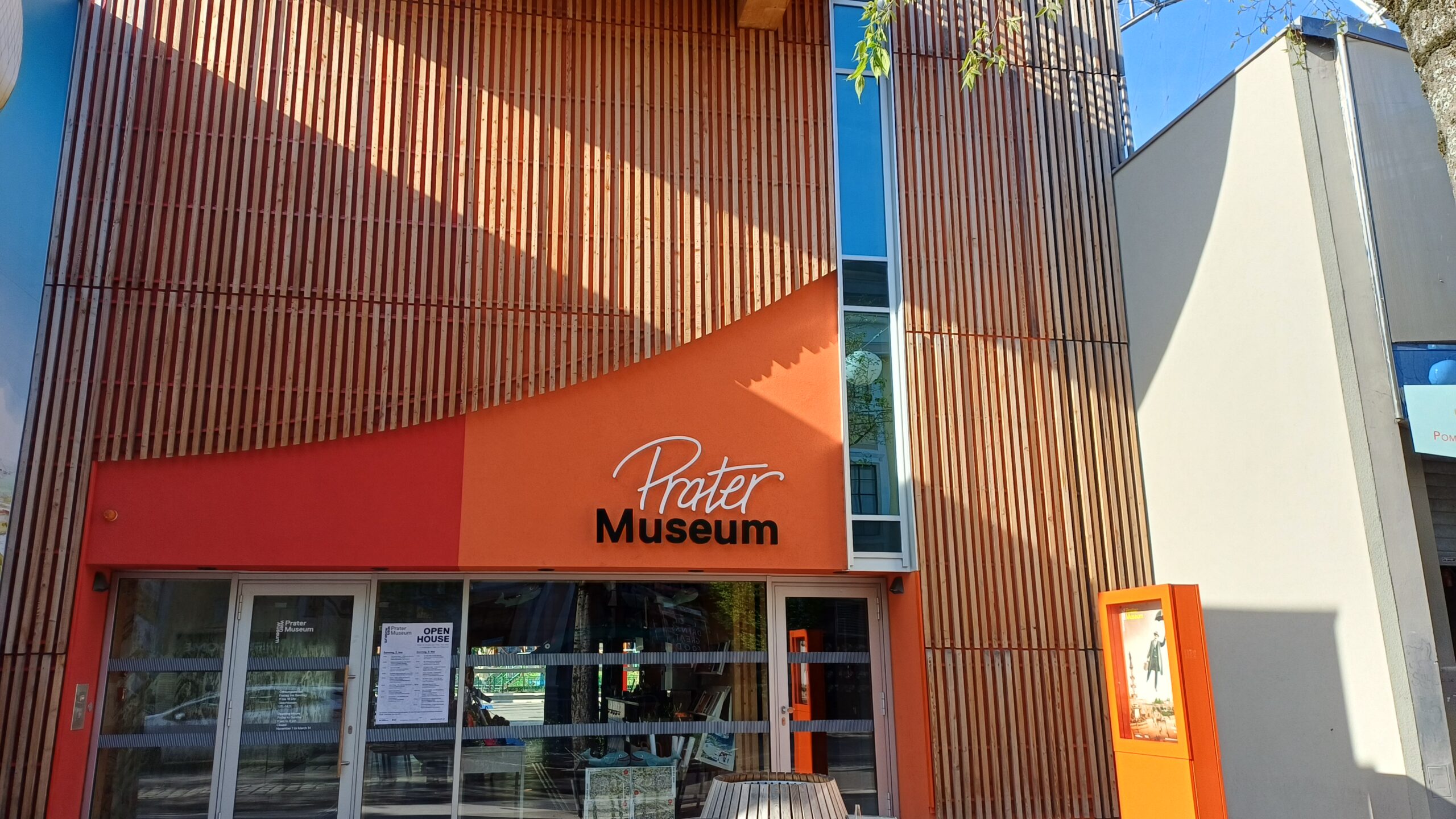 Aufnahme eines Gebäudes mit oranger Fassade und einem verglasten Eingangsbereich, drüber die Aufschrift Prater Museum. rechts vom Eingang ist ein orangener Aufsteller mit einem Plakat