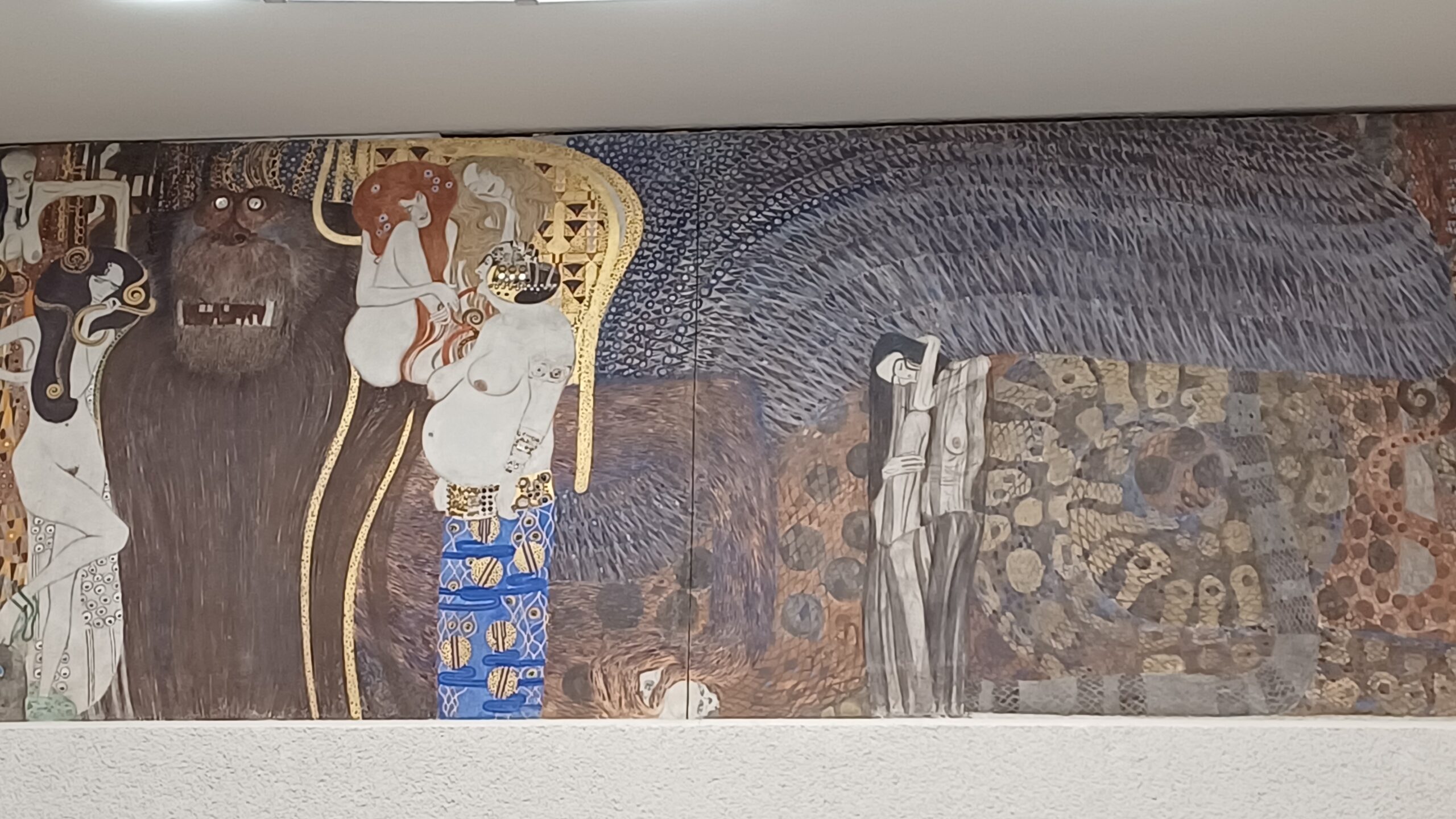 Abbildung des Beethovenfries: Eine braun-blau gemusterte Fläche, auf der Karikaturen von weiblichen Personen und eine monströse Figur in braun abgebildet sind. Im Zentrum ist eine weibliche Person mit langen Haaren in gold und einem goldenen Ornament