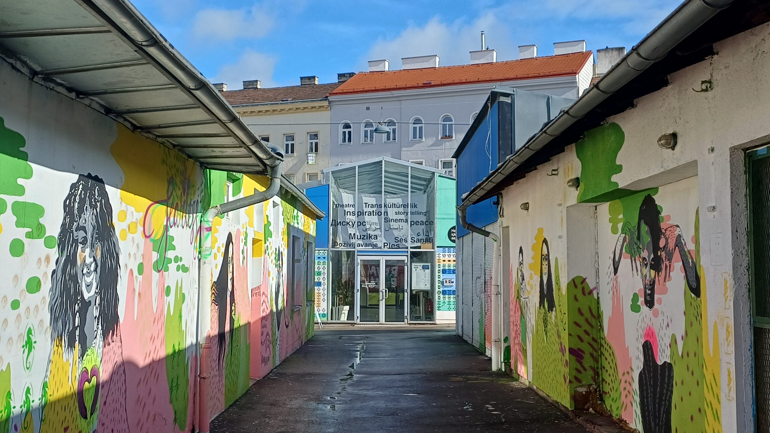 Aufnahme einer Passage, die von zwei Wänden mit grün-gelben Graffitis flakiert sind. Sie führt zu einem Glaseingang, über dem Schriftzüge gedruckt sind