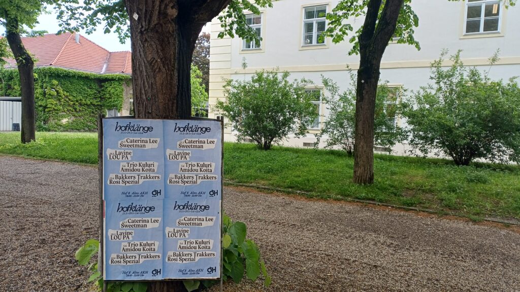 Aufnahme von einem Schotterweg und einer Wiese mit Büschen und Laubbäumen, von denen man die Stämme sieht. Im Zentrum ist ein Plakatständer, auf dem vier hellblaue Plakate angebracht sind, die das Programm der Veranstaltung Hofklänge ankündigen, unten rechts ist das Logo der ÖH