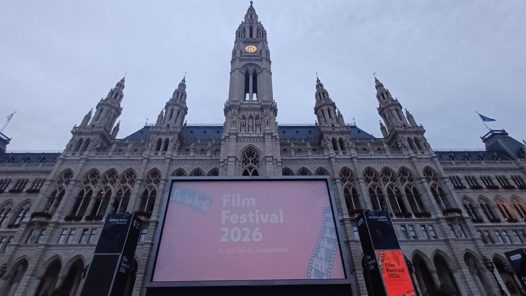Aufnahme des Wiener Rathauses mit seinem spitzen Turm. Davor ist eine Leinwand mit dem weißen Schriftzug Film Festival 2026 auf rotem Hintergrund am Bildschirm